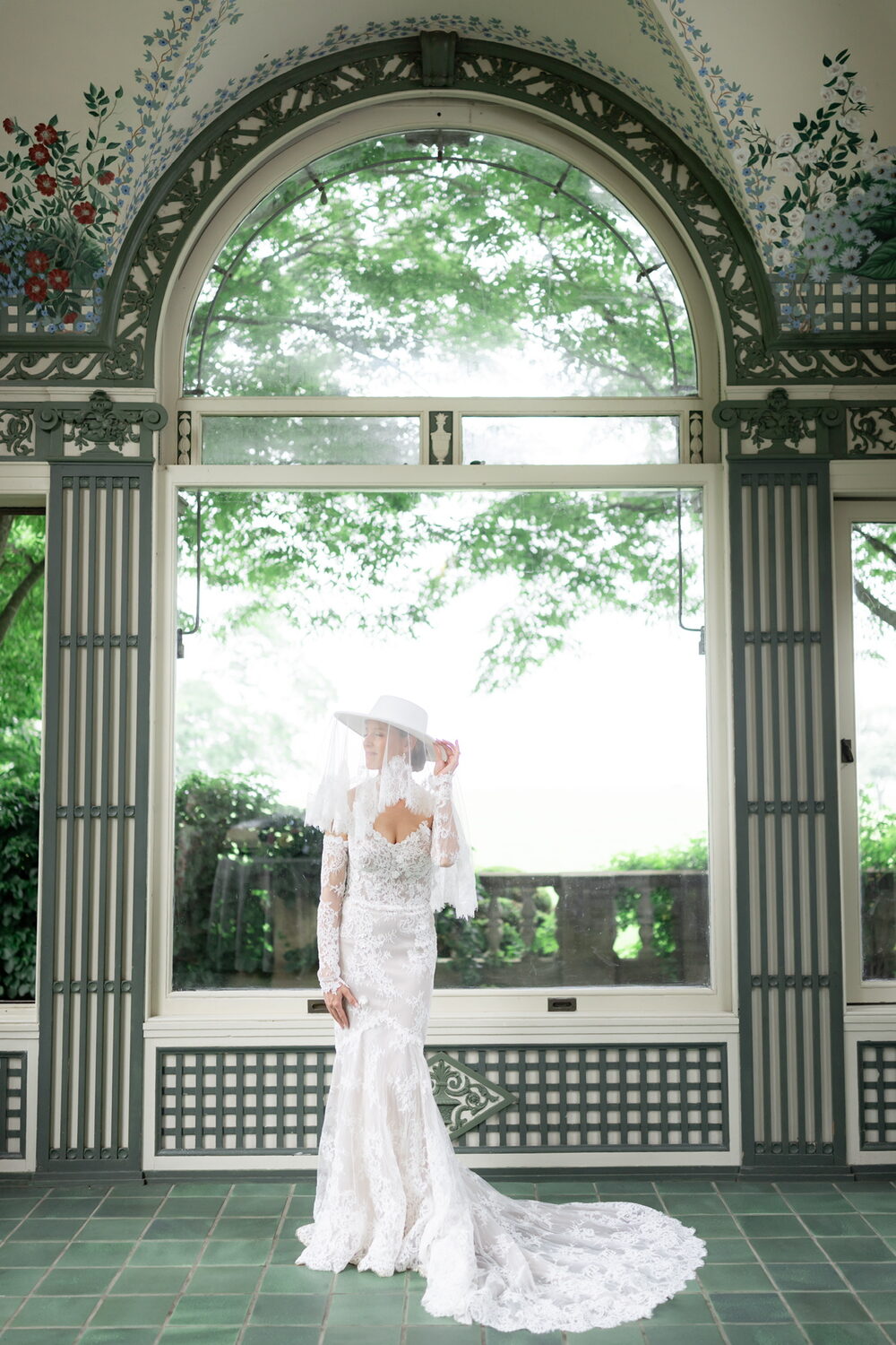 Bride in a lace gown and wide-brim hat beneath a painted garden archway
